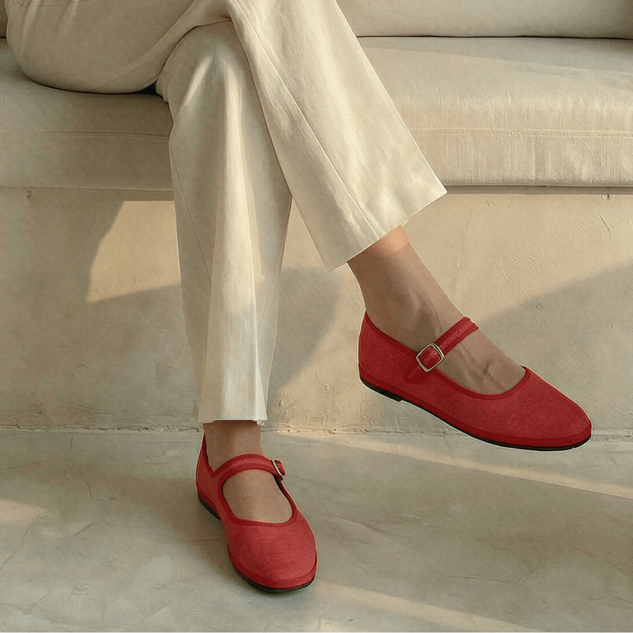 Woman sitting on a beige sofa holding a cup, wearing a white top and pants with red shoes, in a sunlit room with plants.